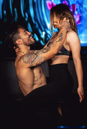 Muscular, shirtless tattooed man grinning as he lifts a woman in a sports bra and high-waisted leggings on a neon-lit dance floor with colorful LED lights.