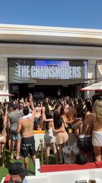 Daytime pool party crowd in swimwear cheering and filming phones at an outdoor stage with a giant 'THE CHAINSMOKERS' marquee on the screen