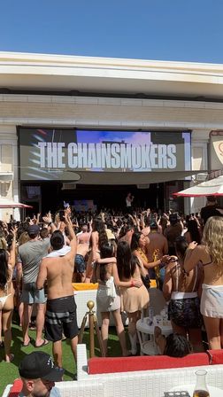 Daytime pool party crowd in swimwear cheering and filming phones at an outdoor stage with a giant 'THE CHAINSMOKERS' marquee on the screen