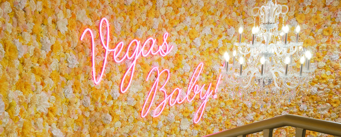 Pink neon "Vegas Baby!" sign on a yellow-and-white floral wall with a sparkling crystal chandelier and a staircase railing in a Las Vegas–style interior