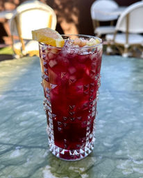 Vibrant deep-red iced beverage in a cut-glass highball with crushed ice, diced fruit and an orange wedge, sitting on a green marble patio table at an outdoor café.