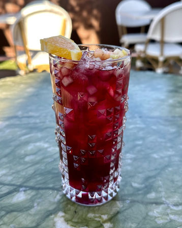 Vibrant deep-red iced beverage in a cut-glass highball with crushed ice, diced fruit and an orange wedge, sitting on a green marble patio table at an outdoor café.