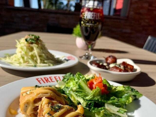 Sunlit outdoor restaurant table with crispy rolled appetizers drizzled in sauce beside a fresh green salad, a bowl of glazed bites, shredded cabbage slaw, and a glass of dark craft beer.