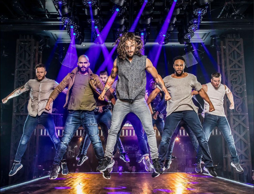 Six male dancers in casual streetwear jumping in unison on a theater stage under purple spotlights and rigging, high-energy urban choreography.
