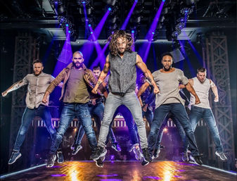 Six male dancers in casual streetwear jumping in unison on a theater stage under purple spotlights and rigging, high-energy urban choreography.