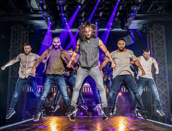Six male dancers in casual streetwear jumping in unison on a theater stage under purple spotlights and rigging, high-energy urban choreography.