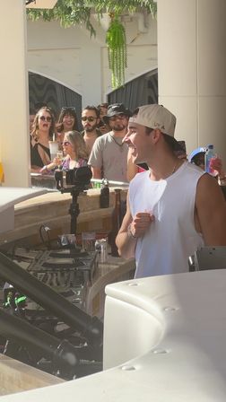 Pool party DJ smiling at an outdoor poolside booth with turntables, camera rig and a cheering crowd holding drinks.
