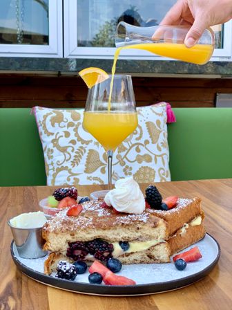 Brunch scene: stuffed French toast with custard and fresh berries, whipped cream and butter, plus a mimosa being poured on a sunny patio