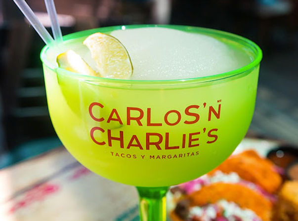 Oversized neon green margarita cup filled with a frozen lime cocktail, lime wedge and two straws, with tacos blurred in the background