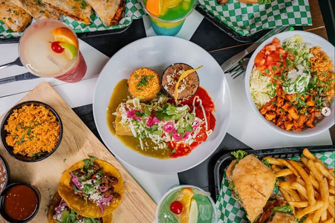 Colorful Mexican food spread on a restaurant table: enchiladas with red and green sauces, Mexican rice and refried beans, tacos, a burrito bowl, a sandwich with fries, and bright tropical cocktails.