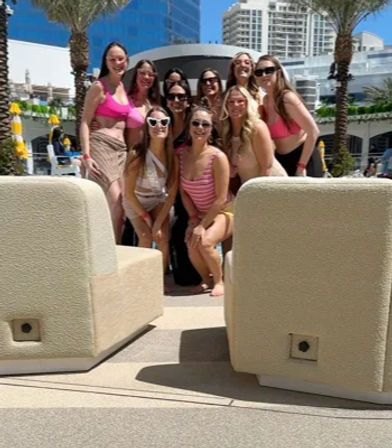 Cheerful group of women in colorful swimsuits posing poolside on a sunny urban resort deck with palm trees, lounge seating and high-rise buildings in the background