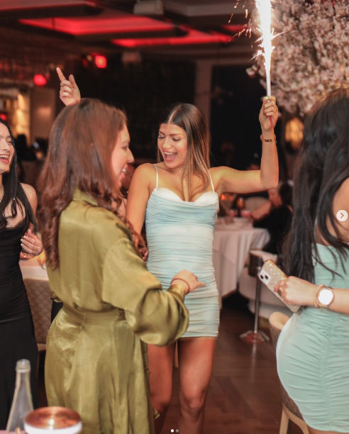 Women celebrating in a dimly lit restaurant lounge — a woman in a light blue cocktail dress holds a lit sparkler while friends in party dresses laugh and dance around a dinner table.