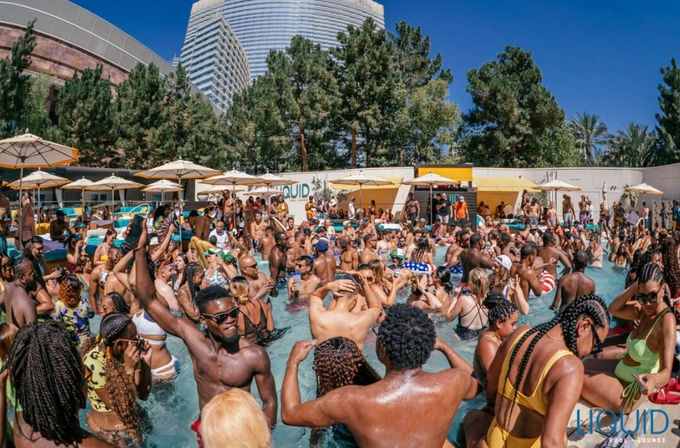 Vibrant outdoor hotel pool party with a large diverse crowd socializing and dancing in the water, yellow umbrellas and lounge chairs, city high-rise backdrop on a sunny day.