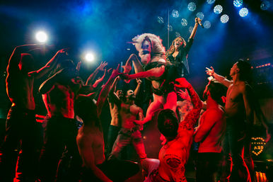 Performer with microphone riding a unicorn prop above a cheering crowd of shirtless dancers on a neon-lit nightclub stage with dramatic spotlights