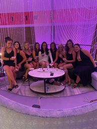 Group of women in cocktail dresses sharing drinks at a curved VIP booth in a purple-lit upscale cocktail lounge, round table with martinis and snacks and a decorative beaded curtain backdrop — girls’ night out vibe.