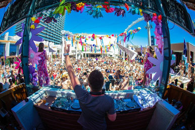 DJ performing from a booth at a sunny outdoor pool party, view over a packed crowd of people in swimsuits cheering and taking photos amid colorful streamers, oversized paper flowers and dancers on stage at a resort-style venue.