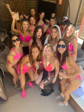 Group of women in bright pink bikinis and cover-ups posing in a sunlit pool cabana, holding drinks with one wearing a white bridal veil — lively daytime bachelorette-style pool party vibe.