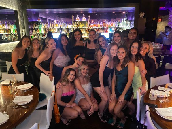 Group of about sixteen smiling women dressed up for a girls' night out, posing in front of a backlit liquor display at an upscale downtown lounge with cocktail tables and menus visible.