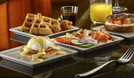 Colorful restaurant brunch spread on a glossy table: eggs benedict with hollandaise, waffles with syrup and fried bites, smoked salmon with cucumber and red onion, poppy-seed bagel with cream cheese, and a glass of orange juice.