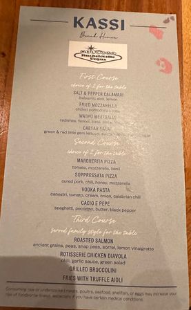 Close-up of a printed Italian-American restaurant menu on a wooden table listing first, second and third courses — salt & pepper calamari, fried mozzarella, wagyu meatballs, margherita and soppressata pizzas, vodka pasta, cacio e pepe, roasted salmon, rotisserie chicken diavola, grilled broccolini and fries with truffle aioli — lipstick marks on the top corner.