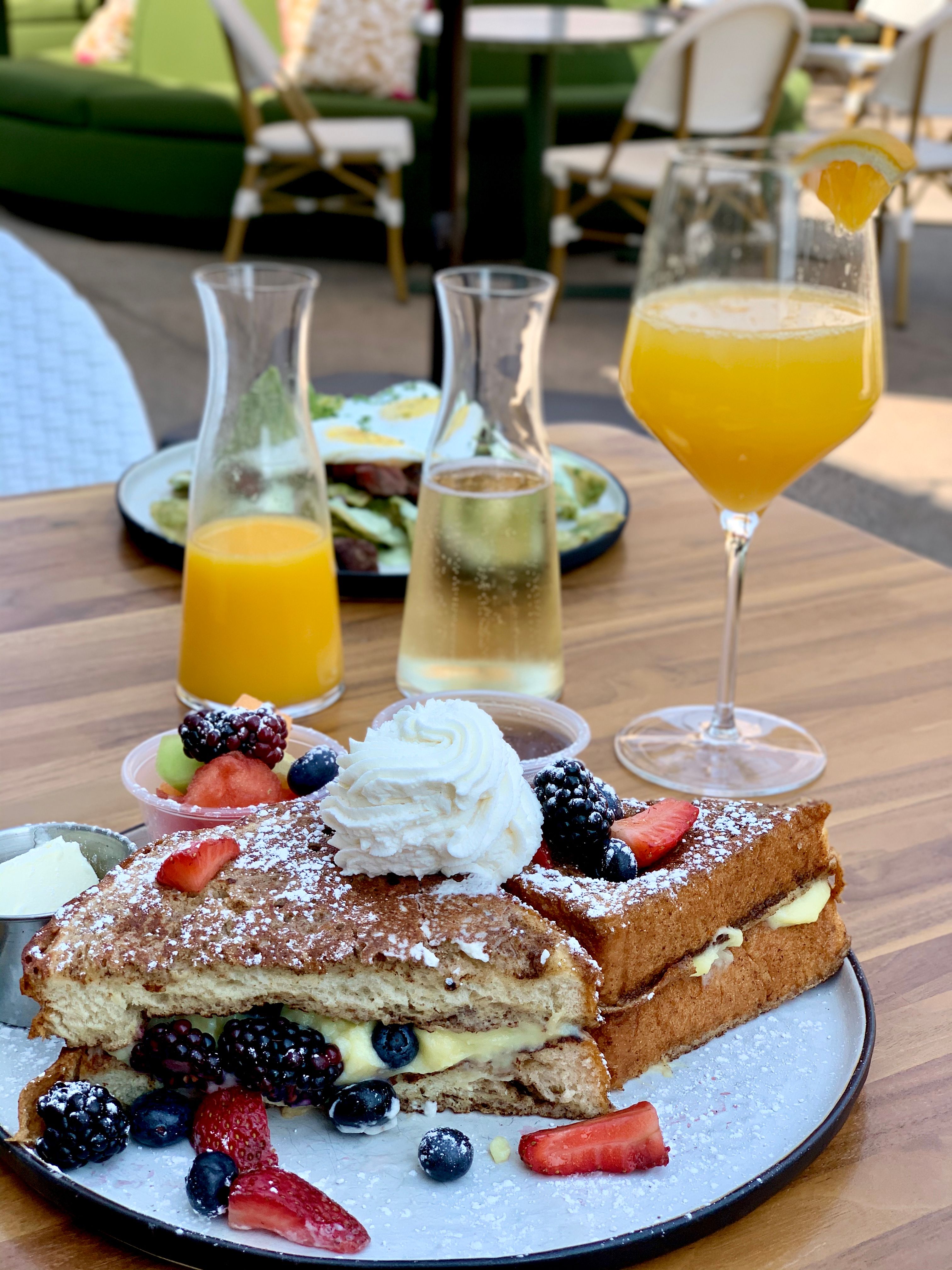 Stuffed French toast with custard, fresh berries, whipped cream and powdered sugar on a plate, served on a wooden patio table with fruit cups, carafes of orange juice and sparkling wine and a mimosa — sunny outdoor brunch scene.