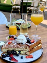 Stuffed French toast with custard, fresh berries, whipped cream and powdered sugar on a plate, served on a wooden patio table with fruit cups, carafes of orange juice and sparkling wine and a mimosa — sunny outdoor brunch scene.