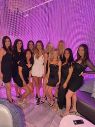 Group of women in black and white cocktail dresses smiling and posing in front of a purple LED bead curtain inside a trendy lounge, evening night-out vibe.