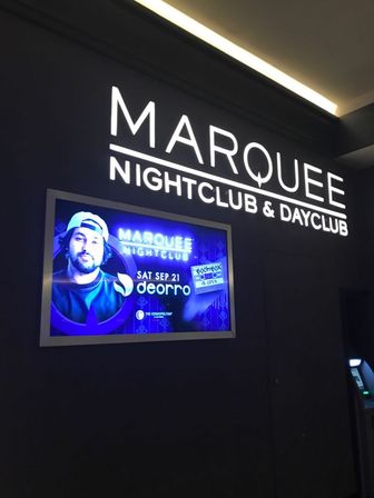 Bright indoor nightclub and dayclub entrance with a bold illuminated wall sign above a framed purple-blue digital poster promoting a DJ night