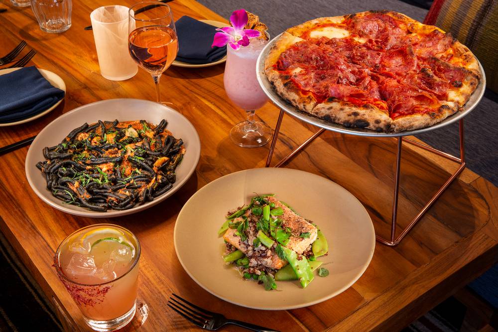 Cozy wood-table restaurant spread: Neapolitan pepperoni pizza on a metal stand, black squid-ink pasta, seared salmon with snap peas, plus rosé and colorful cocktails (one garnished with a pink orchid).