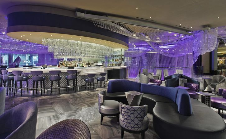 Glamorous modern lounge bar interior with a curved marble bar, cascading crystal chandeliers, purple ambient lighting and plush curved seating.