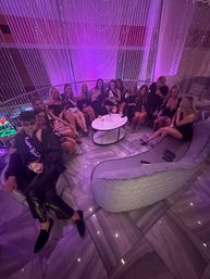 Group of women in black dresses lounging on a curved sofa in a purple-lit upscale lounge with string-light curtains, marble floor and drinks on a round table — lively bachelorette party vibe