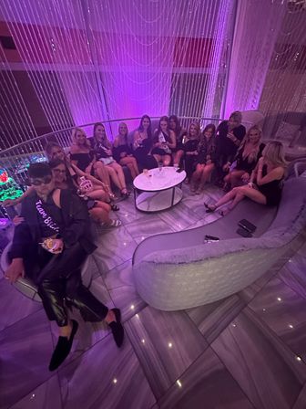 Group of women in black dresses lounging on a curved sofa in a purple-lit upscale lounge with string-light curtains, marble floor and drinks on a round table — lively bachelorette party vibe