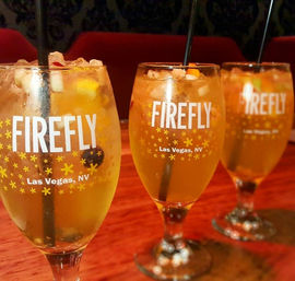 Three chilled, fruit-garnished cocktails in stemmed glasses on a wooden bar; glasses show 'Las Vegas, NV' text with star motifs, filled with ice and black straws.