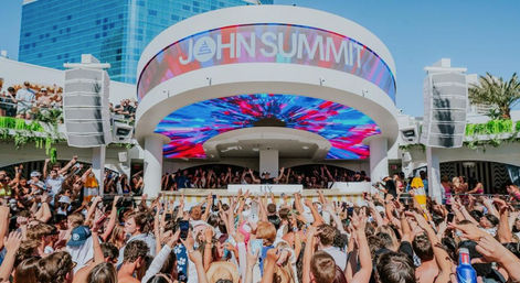 Daytime outdoor pool party at a packed rooftop dayclub, crowd with hands raised toward a DJ booth under a vivid multicolored LED canopy and high-rise hotel in the background.