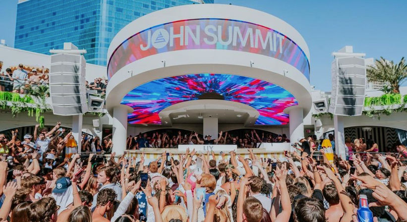 Daytime outdoor pool party at a packed rooftop dayclub, crowd with hands raised toward a DJ booth under a vivid multicolored LED canopy and high-rise hotel in the background.