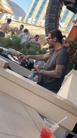 Tattooed DJ with a man bun mixing on decks at a sunny outdoor pool club, shirtless party crowd behind him and a red cocktail in the foreground.