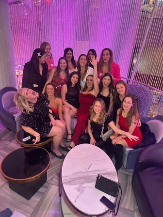 Group of women in cocktail dresses posing and laughing on lounge seating with a pink beaded curtain backdrop — festive girls' night out in an upscale lounge