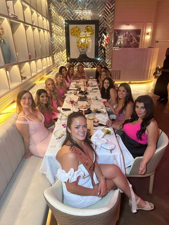 Cheerful bachelorette party of women in pink and white dresses seated at a long restaurant table with cocktails and plates, gold-accented mirrored wall and framed art in a chic, modern dining room.