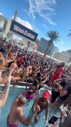High-energy daytime Las Vegas pool party with a packed crowd in swimwear, a performer on the pool deck, palm trees and Strip hotels under a bright blue sky.
