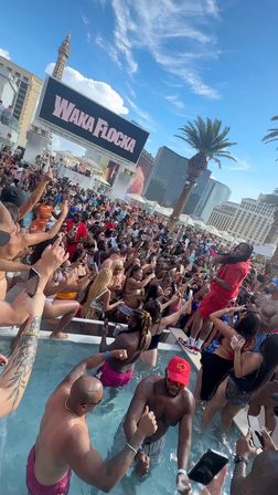 High-energy daytime Las Vegas pool party with a packed crowd in swimwear, a performer on the pool deck, palm trees and Strip hotels under a bright blue sky.
