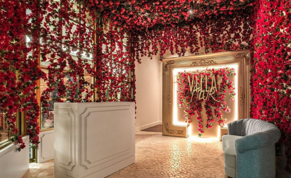 Indoor boutique lobby overflowing with hanging red roses, a white reception desk, ornate illuminated gold photo frame with floral backdrop, and a curved gray sofa on textured tile floor — a romantic, Instagrammable entrance.