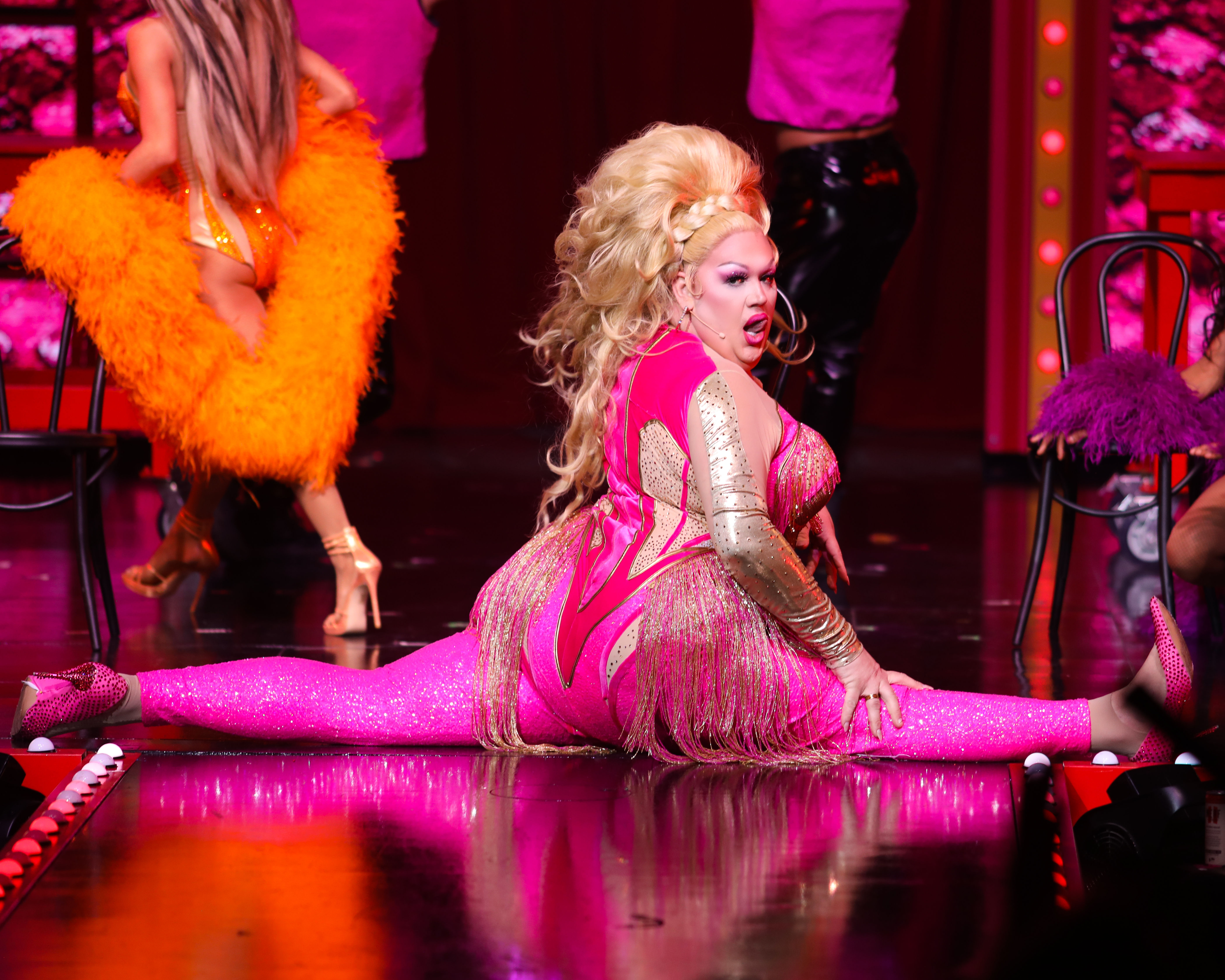 Drag performer in a glittering hot-pink fringed costume doing a full split on a theatrical stage during a vibrant cabaret-style show with feathered dancers and colorful lighting.