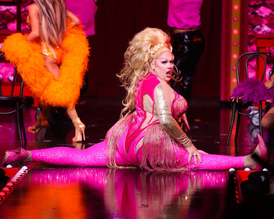 Drag performer in a glittering hot-pink fringed costume doing a full split on a theatrical stage during a vibrant cabaret-style show with feathered dancers and colorful lighting.