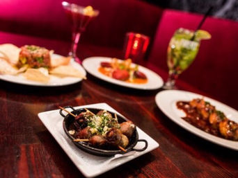 Skillet of parmesan-topped meatballs on a wood table surrounded by tapas-style small plates, colorful cocktails, and a vibrant red restaurant booth