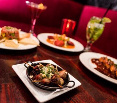 Skillet of parmesan-topped meatballs on a wood table surrounded by tapas-style small plates, colorful cocktails, and a vibrant red restaurant booth