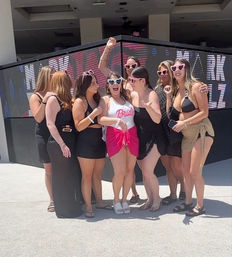 Sunny poolside bachelorette party group photo — bride in a white 'Bride' swimsuit and pink sarong wearing heart-shaped sunglasses surrounded by friends in black swimwear and playful shades at an outdoor dayclub.