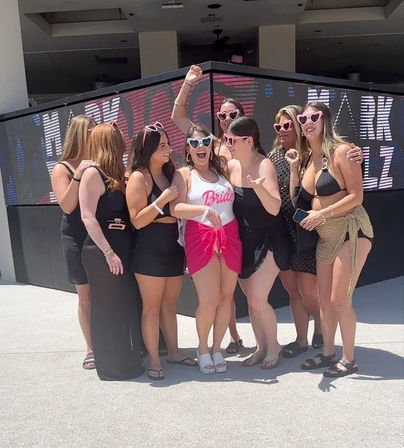 Sunny poolside bachelorette party group photo — bride in a white 'Bride' swimsuit and pink sarong wearing heart-shaped sunglasses surrounded by friends in black swimwear and playful shades at an outdoor dayclub.