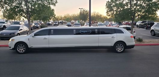 Side profile of a sleek white stretch limousine parked curbside in a busy parking lot at dusk, framed by trees and rows of cars.