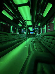 Nightclub-style party bus interior lit in green LEDs with tufted leather bench seats lining the sides, a central dance pole, and illuminated ceiling panels.