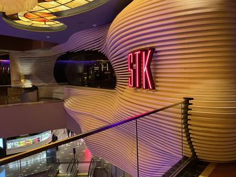 Wavy wood-slat interior wall with glowing red neon letters, glass railing overlooking escalators and a lower concourse, warm ambient lighting and stained-glass ceiling detail.
